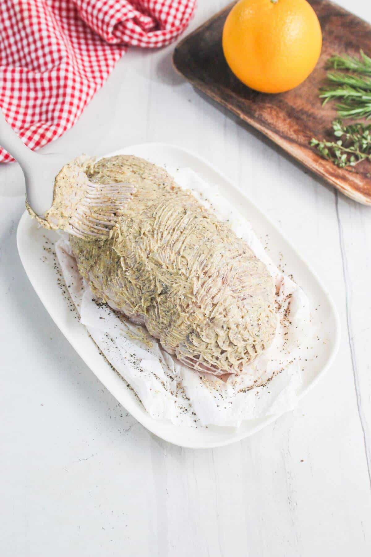 Raw meat coated with a herb and mustard mixture on a white dish, with an orange, herbs, and a red checkered cloth in the background.