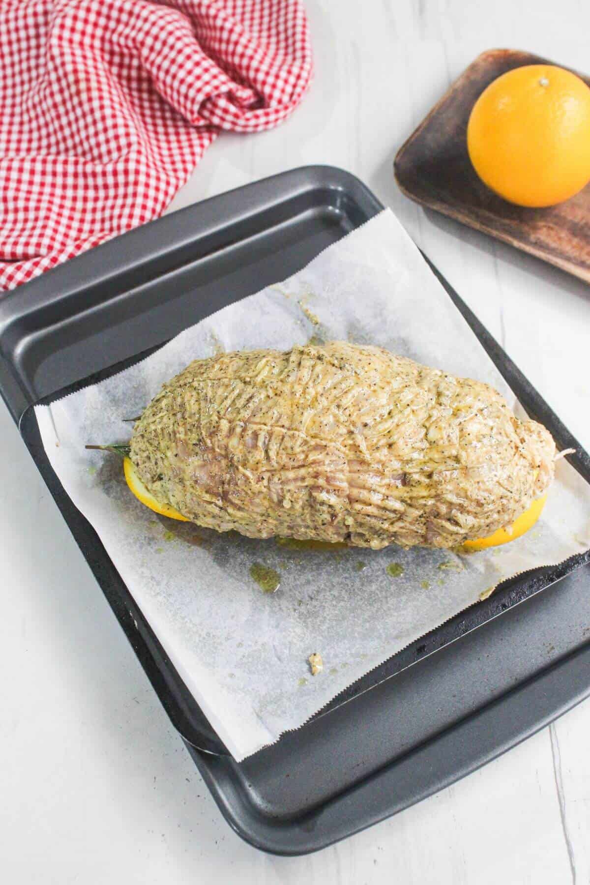 A seasoned, uncooked roast placed on lemon slices sits on a parchment-lined baking tray. An orange and a red checkered cloth are visible in the background.