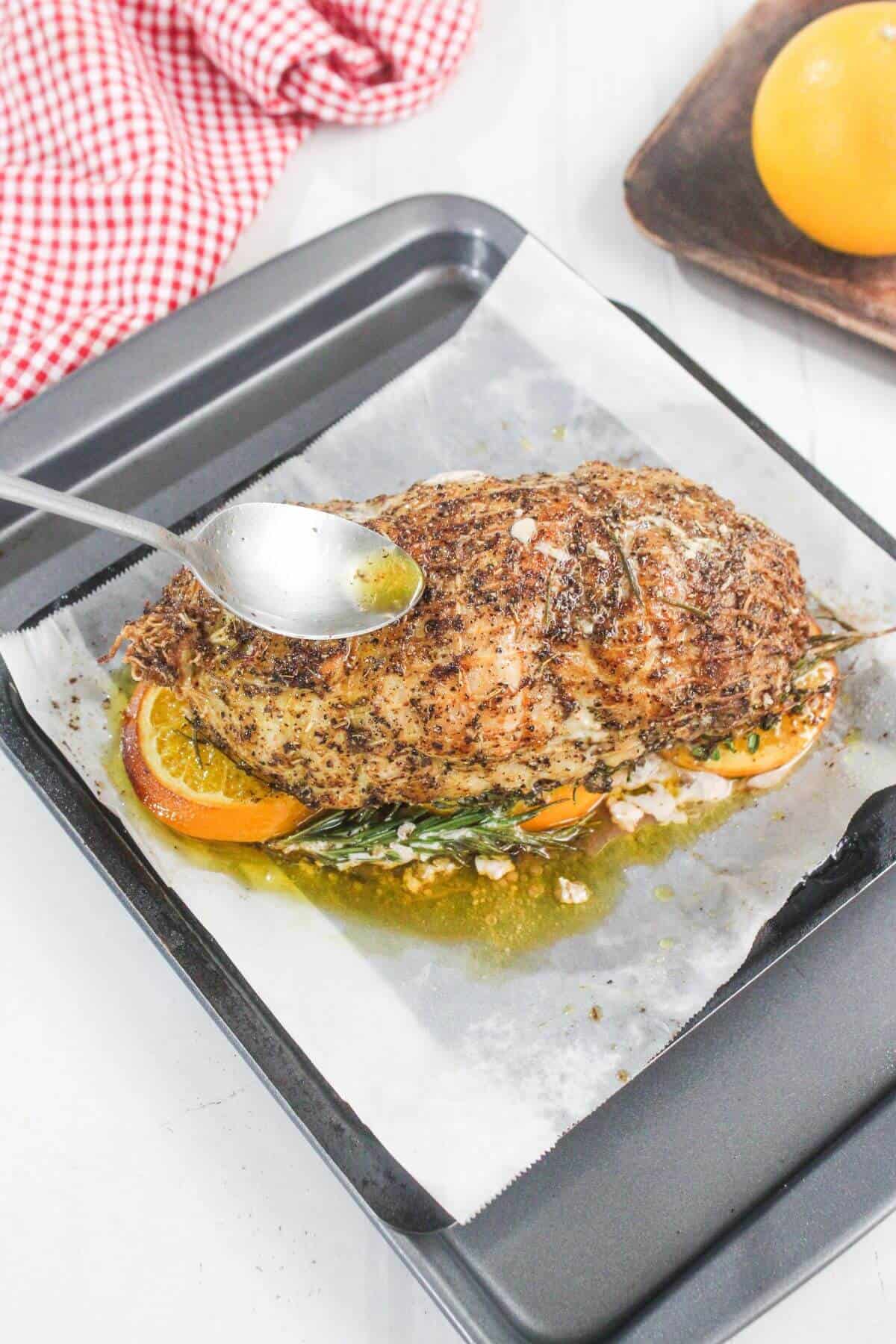 A spoon pours juices over a seasoned, roasted meat atop orange slices and herbs on parchment in a baking tray. A red-checked cloth and an orange are in the background.