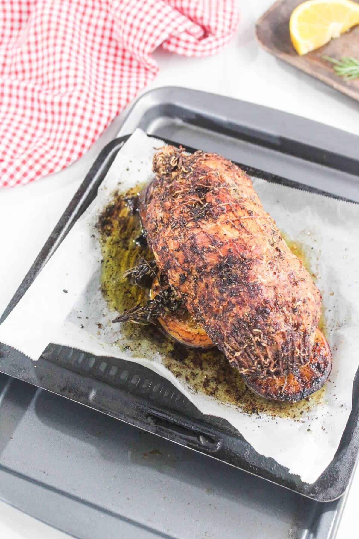 A cooked, herb-crusted roast sits on parchment paper in a baking tray, with roasted juices surrounding it. A red checkered cloth and a lemon wedge are nearby.