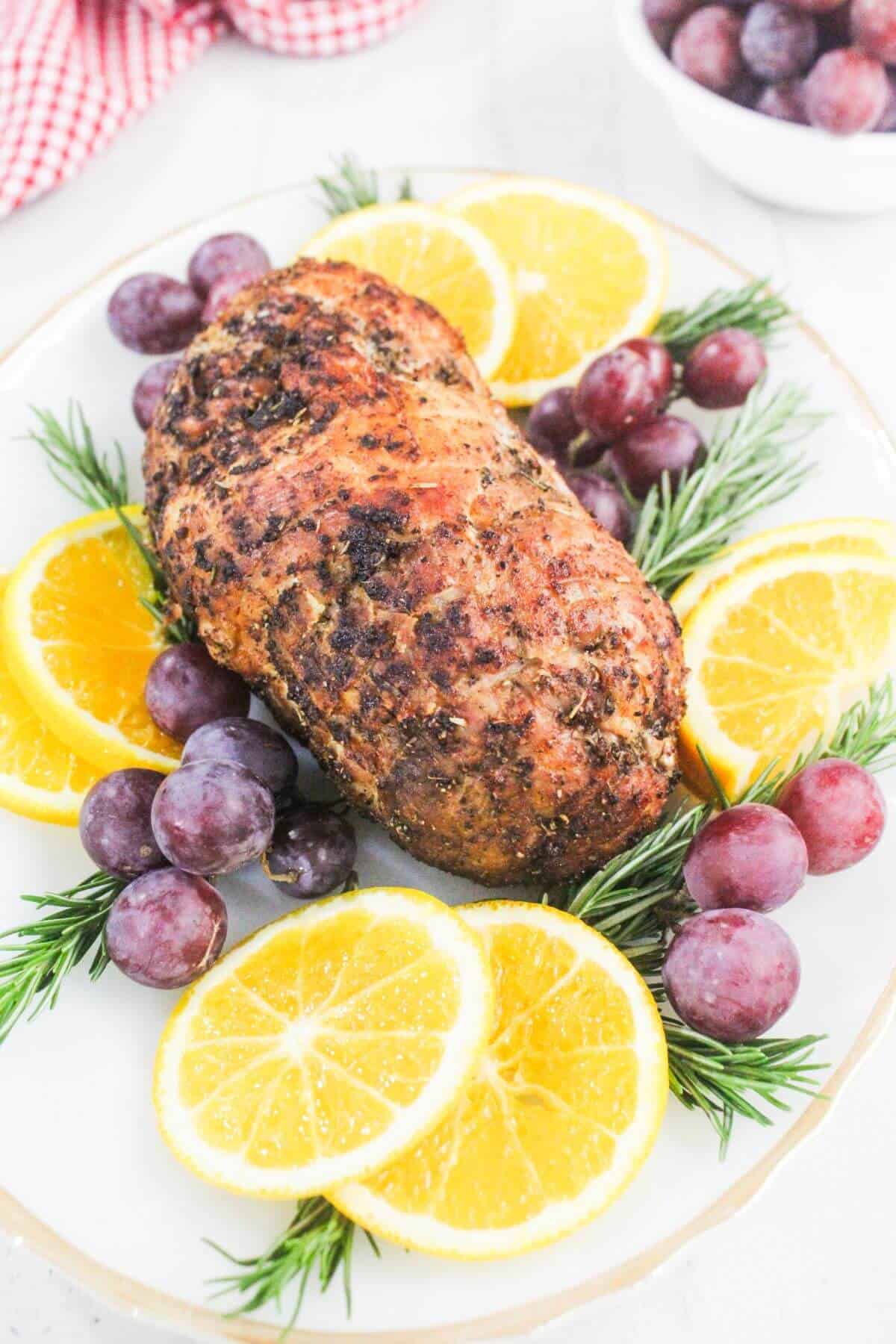 A seasoned roast pork tenderloin on a white plate, garnished with orange slices, red grapes, and sprigs of rosemary.