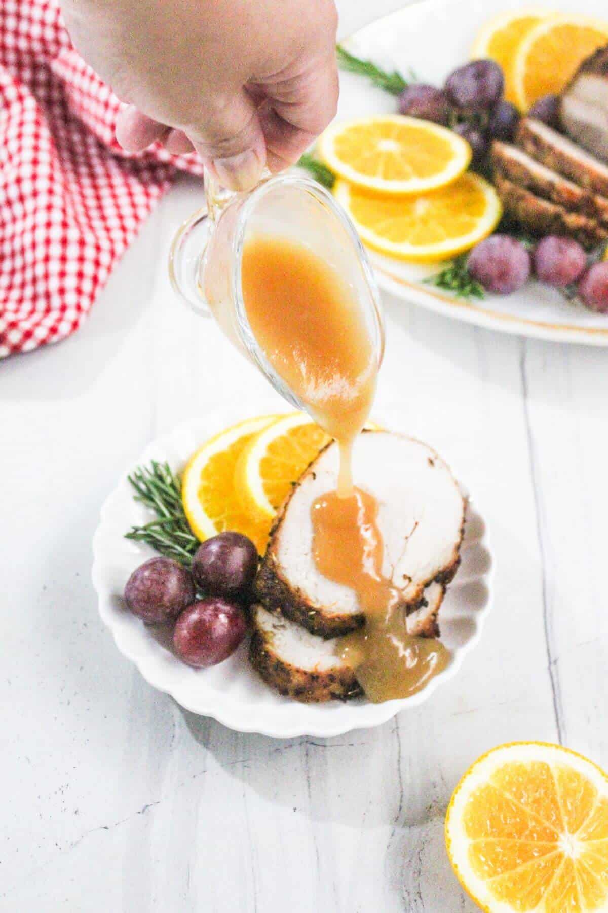 A hand pours gravy over sliced roast pork, served with orange slices, red grapes, and a sprig of rosemary on a white plate.