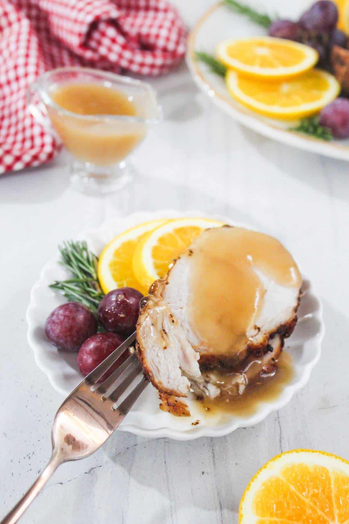 A plate with a slice of roasted turkey topped with gravy, garnished with orange slices, grapes, and rosemary, with a fork and a small gravy dish nearby.
