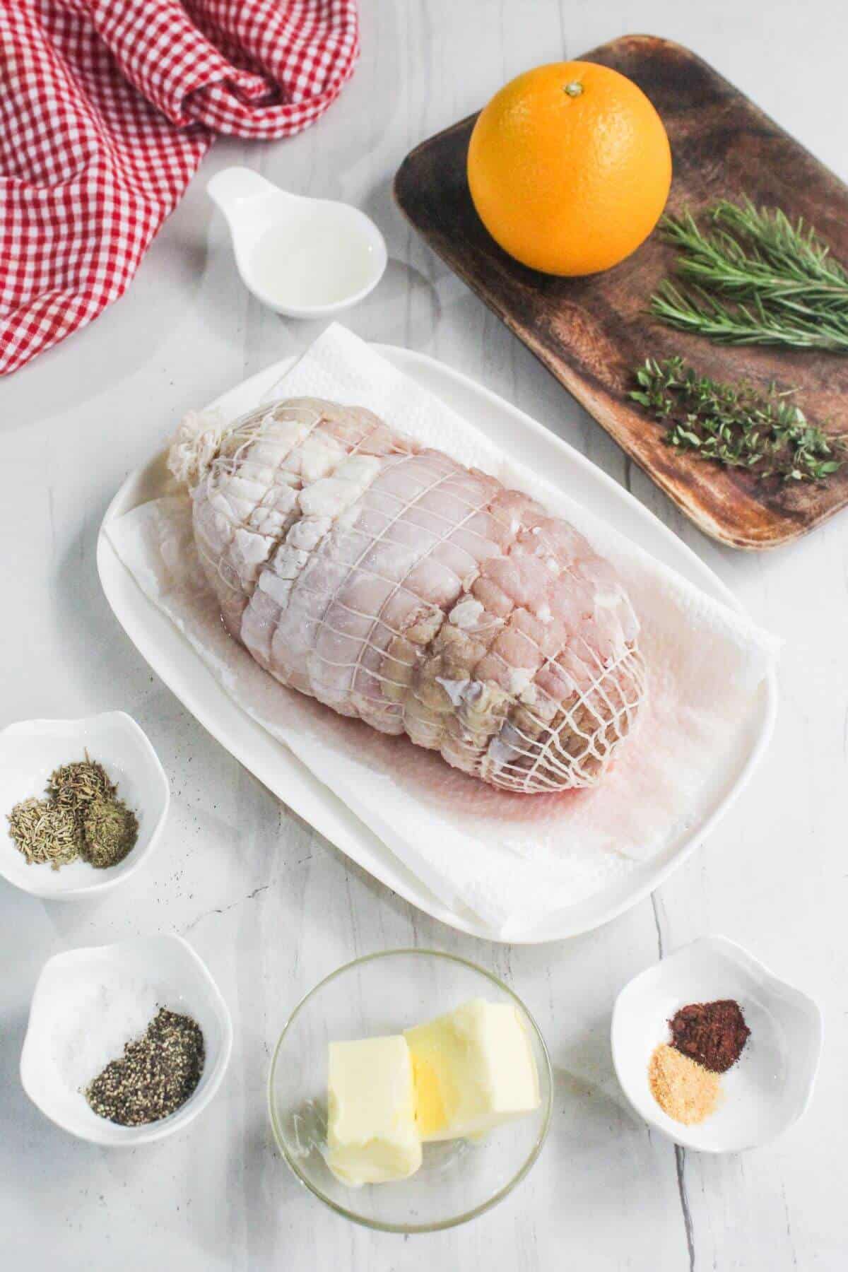 Uncooked boneless turkey roast in a dish, surrounded by butter, spices, herbs, an orange, and a small pitcher of liquid on a white surface.