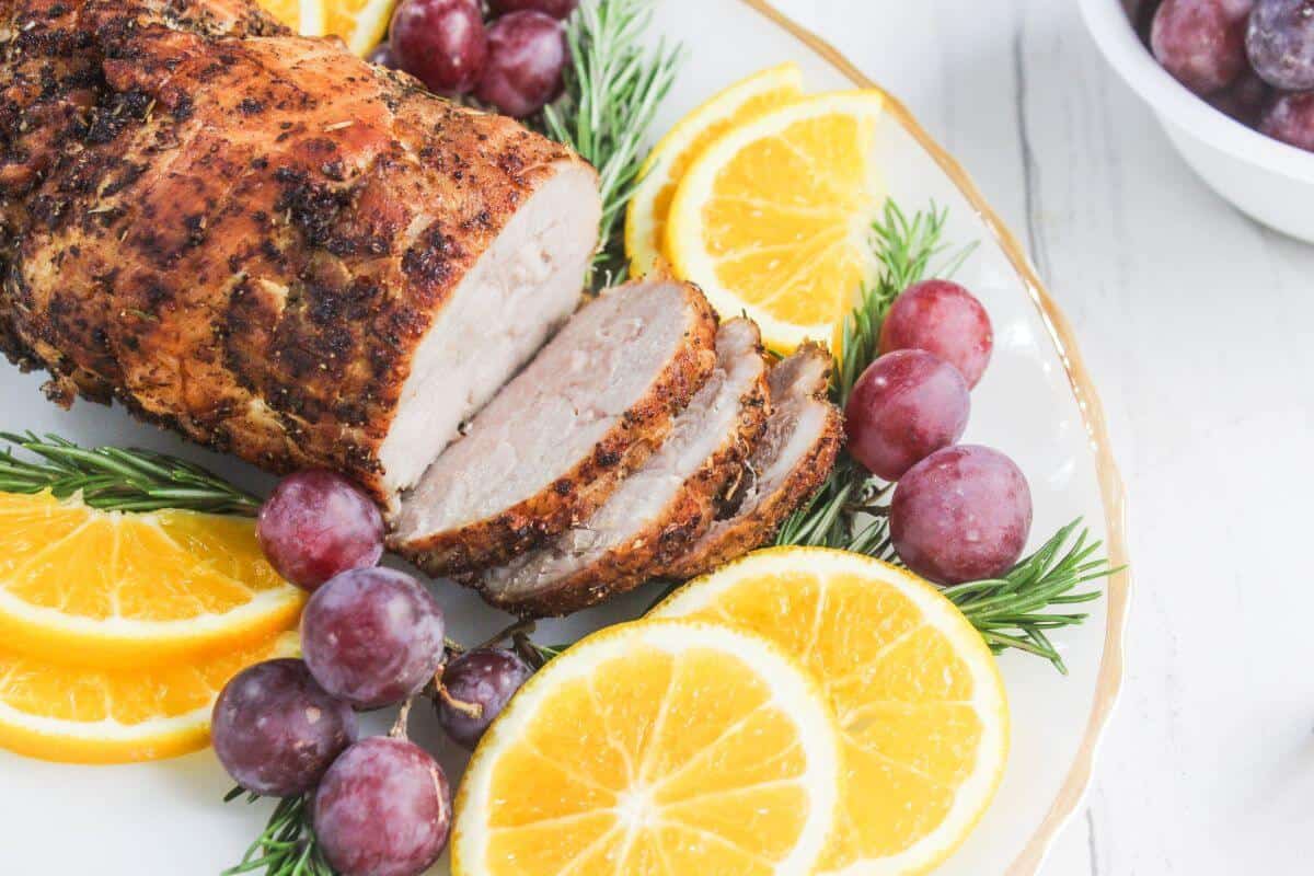 A sliced roasted pork tenderloin on a plate, garnished with orange slices, red grapes, and sprigs of rosemary.