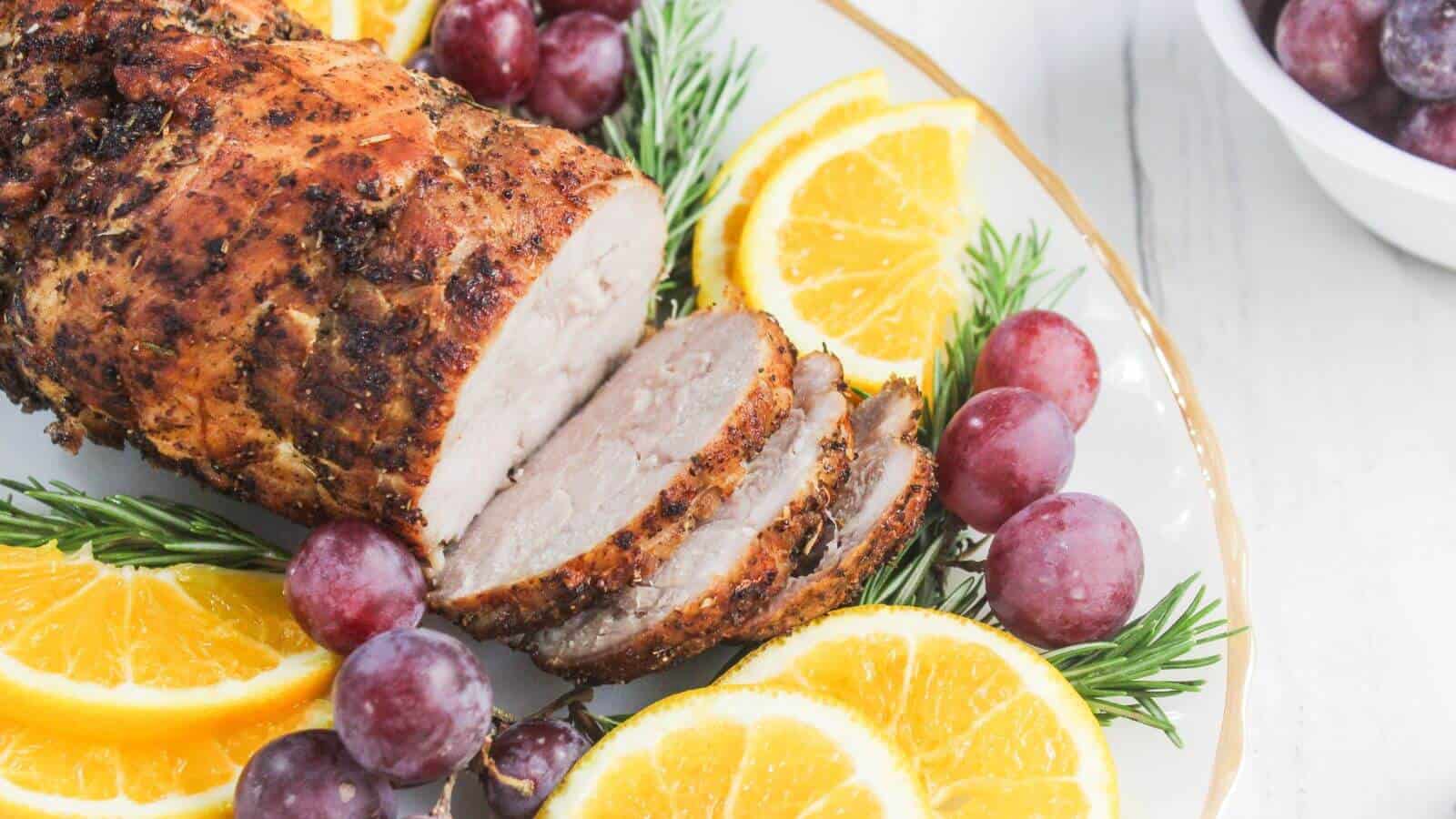 A sliced roasted pork loin garnished with rosemary, orange slices, and red grapes on a white plate, with extra grapes in a bowl nearby.