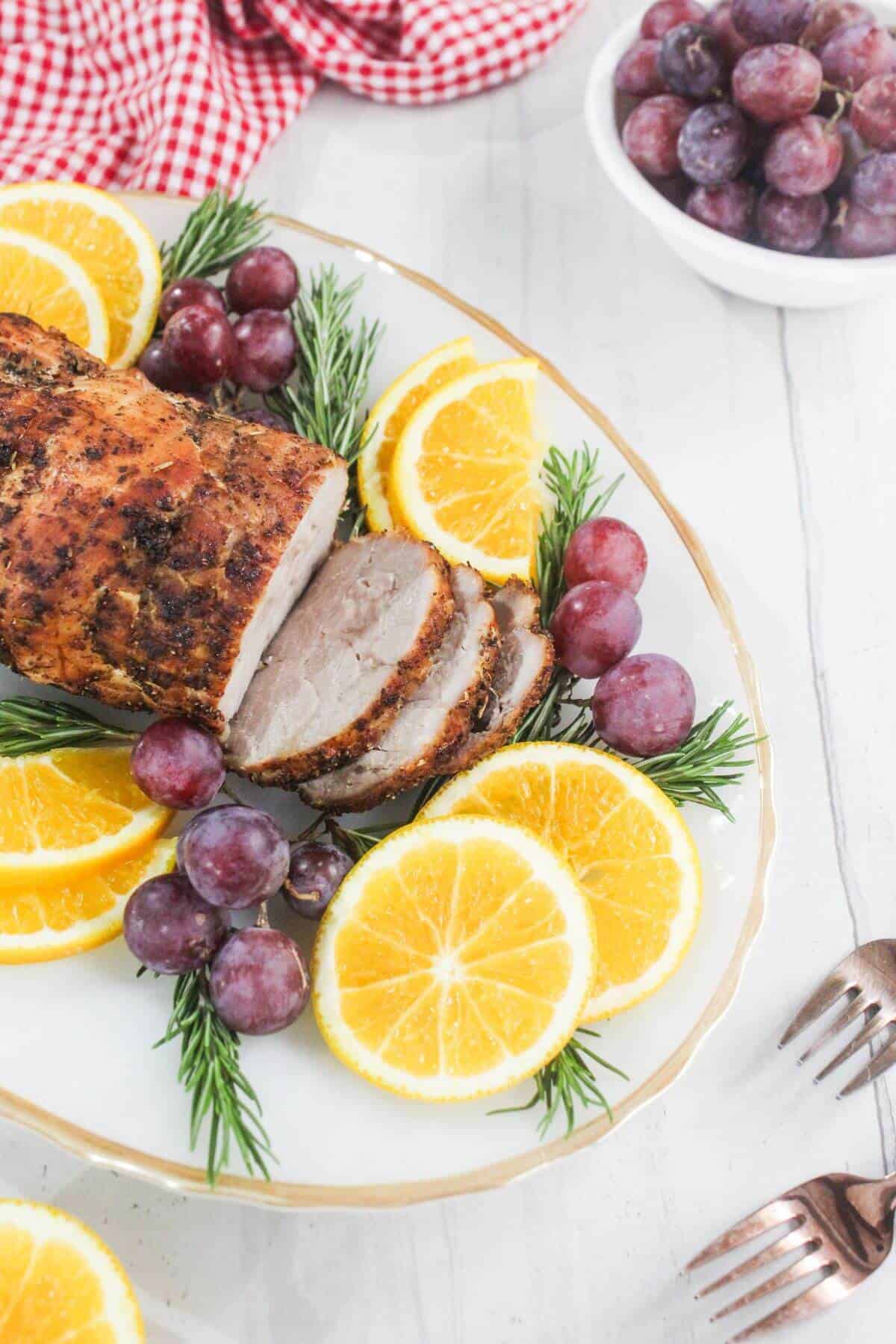 Sliced roasted pork loin garnished with orange slices, red grapes, and rosemary on a white platter, with a bowl of grapes in the background.