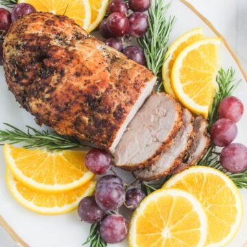 A sliced roasted pork tenderloin garnished with fresh rosemary, orange slices, and red grapes on a white platter.