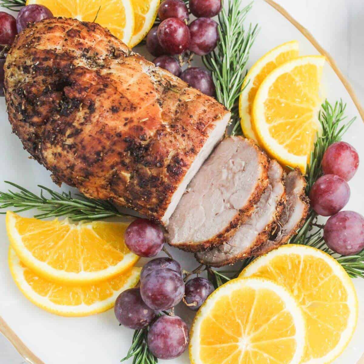 A sliced roasted pork tenderloin garnished with fresh rosemary, orange slices, and red grapes on a white platter.