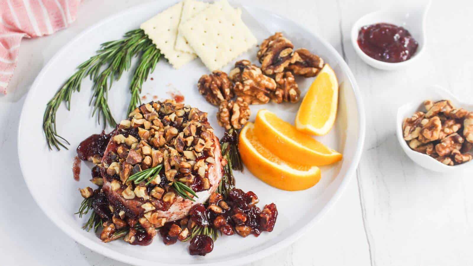 A plate with a cheese round coated in chopped nuts and herbs, surrounded by crackers, walnuts, orange slices, and dollops of dark red jelly.