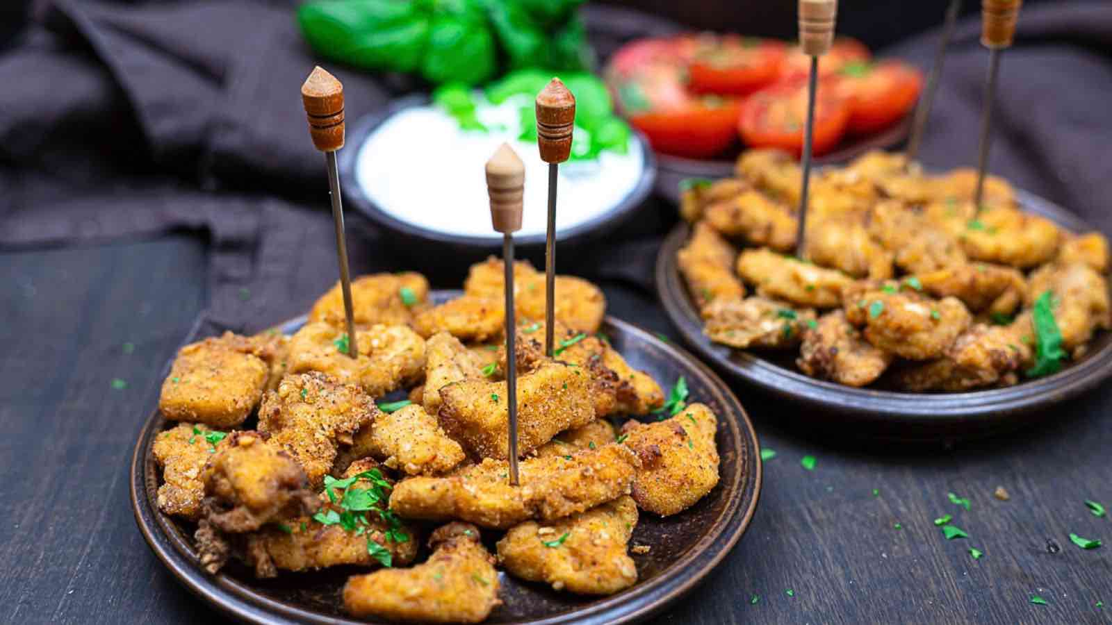 Two plates of breaded and seasoned chicken bites are served with toothpicks, garnished with herbs, and accompanied by dipping sauce and fresh vegetables in the background.