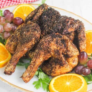 A plate of roasted chicken halves seasoned with herbs, garnished with fresh orange slices, red grapes, and parsley on a white platter.