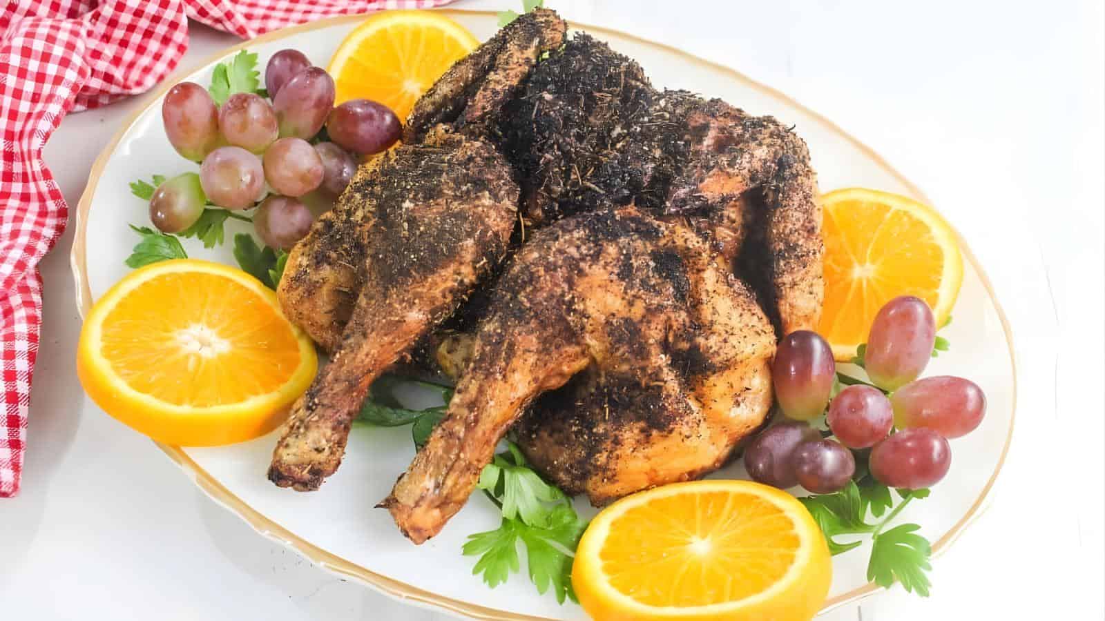 A plate of roasted chicken halves garnished with herbs, surrounded by orange slices, red grapes, and parsley on a white platter.