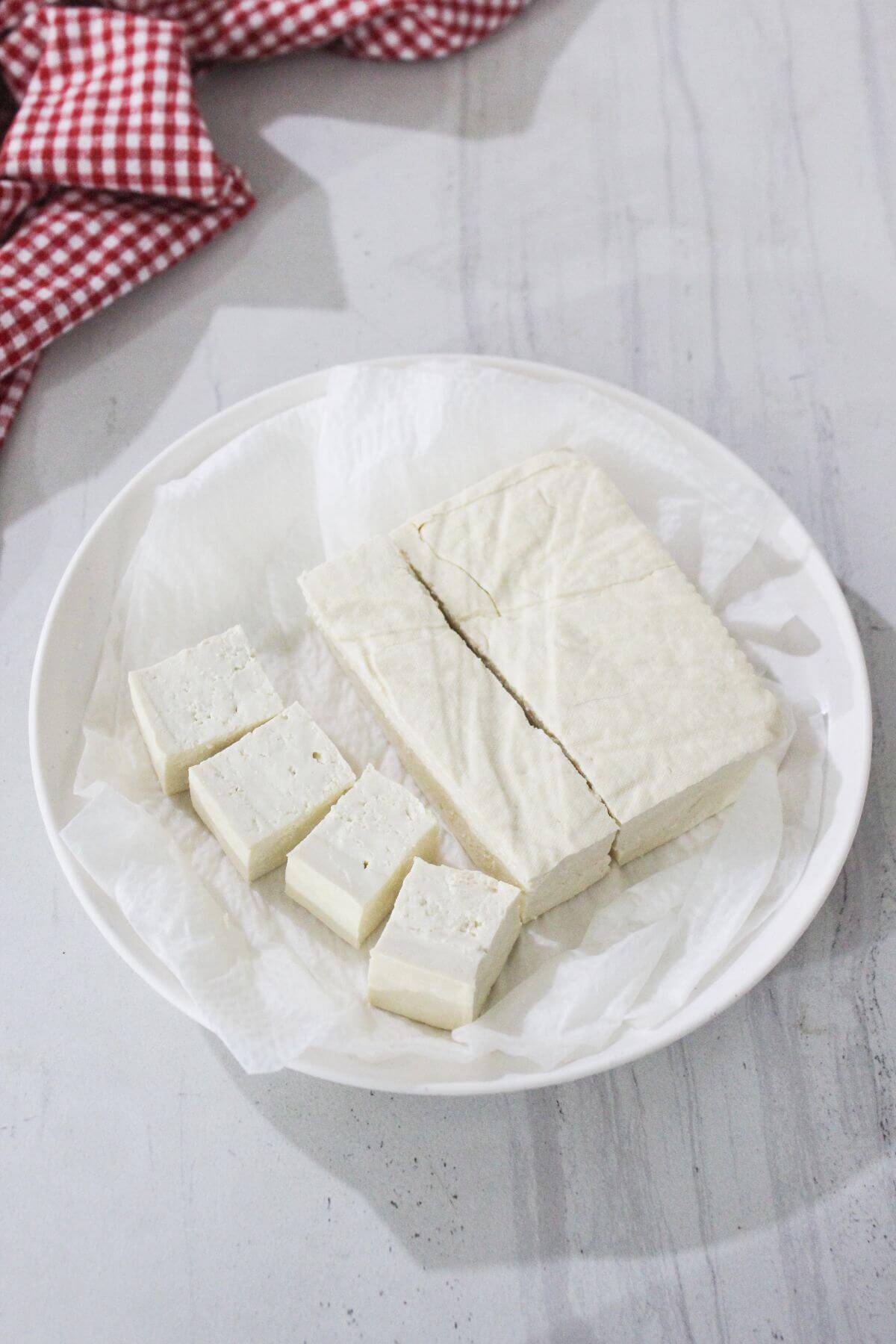 A plate with several pieces of sliced tofu on parchment paper, set on a light-colored surface with a red and white checkered cloth in the background.
