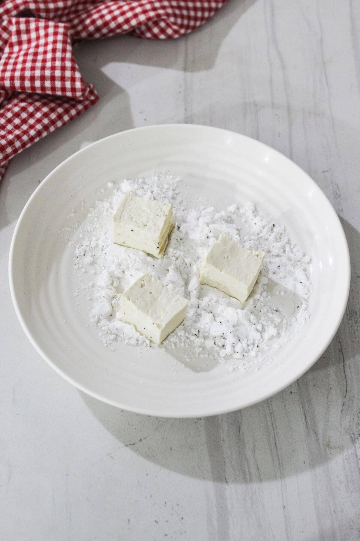 Three square pieces of tofu rest on a white plate dusted with flour, with a red and white checkered cloth in the background.