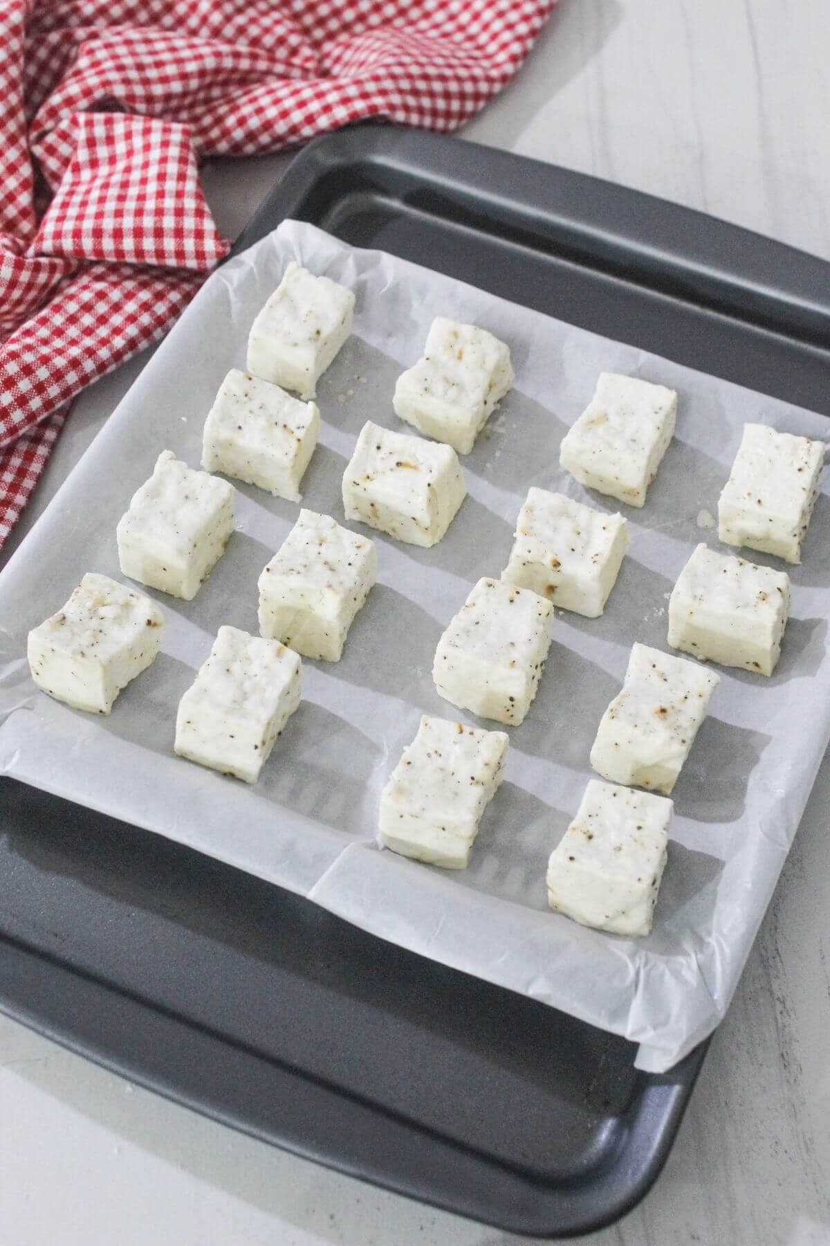 A black baking tray lined with parchment paper holds sixteen evenly spaced cubes of seasoned dough, ready to be baked. A red and white checkered cloth is in the background.