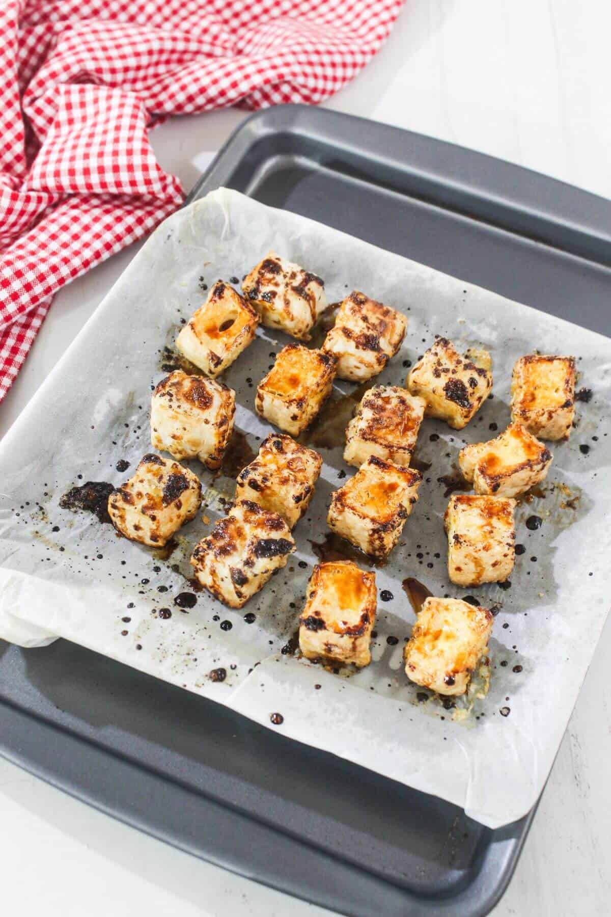 Fifteen browned tofu cubes on parchment-lined baking tray, with a red and white checkered cloth nearby on a white surface.