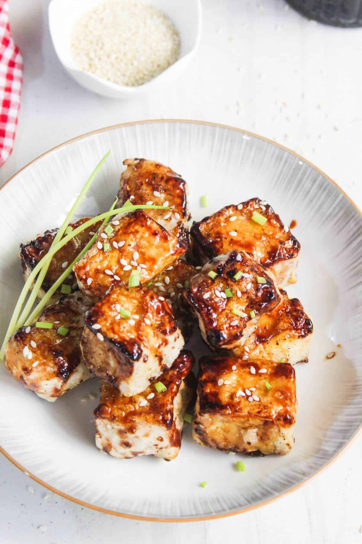 A plate of glazed tofu cubes garnished with sesame seeds and green onions, with a small bowl of sesame seeds in the background.