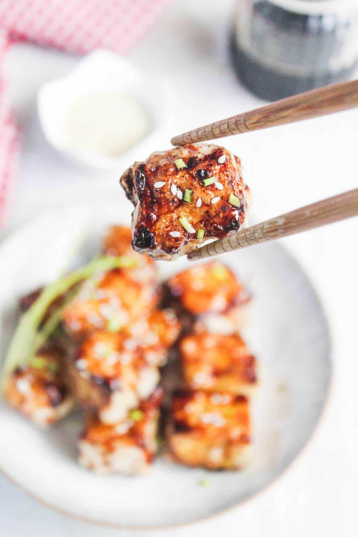 A pair of chopsticks holds a glazed, grilled tofu cube over a plate filled with similar tofu pieces garnished with green onions and sesame seeds.