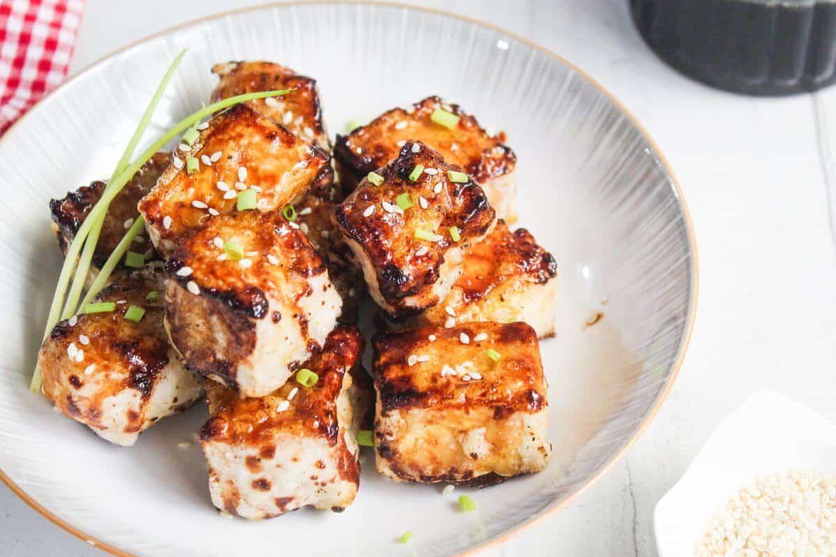 A plate of grilled tofu cubes garnished with sesame seeds and chopped green onions, with a sprig of chive on top.