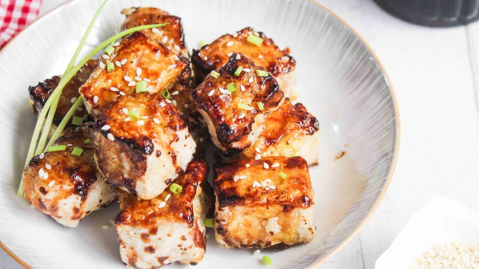 A plate of browned tofu cubes garnished with sesame seeds, chopped green onions, and chives, served in a white bowl.
