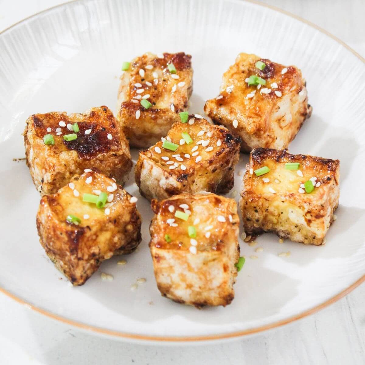 Seven pieces of grilled tofu garnished with chopped green onions and sesame seeds are arranged on a white plate.
