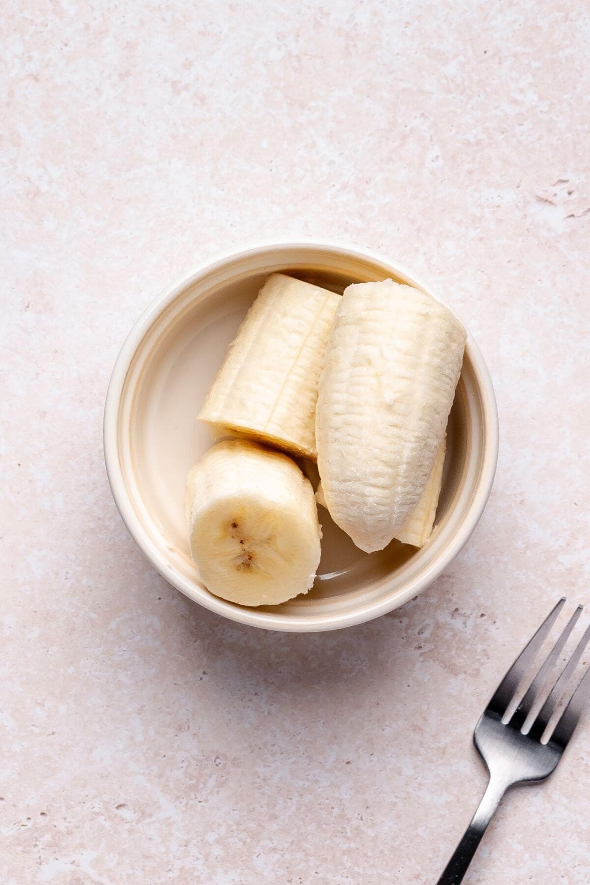 A peeled banana cut into three pieces in a small bowl, with a fork placed nearby on a light-colored surface.