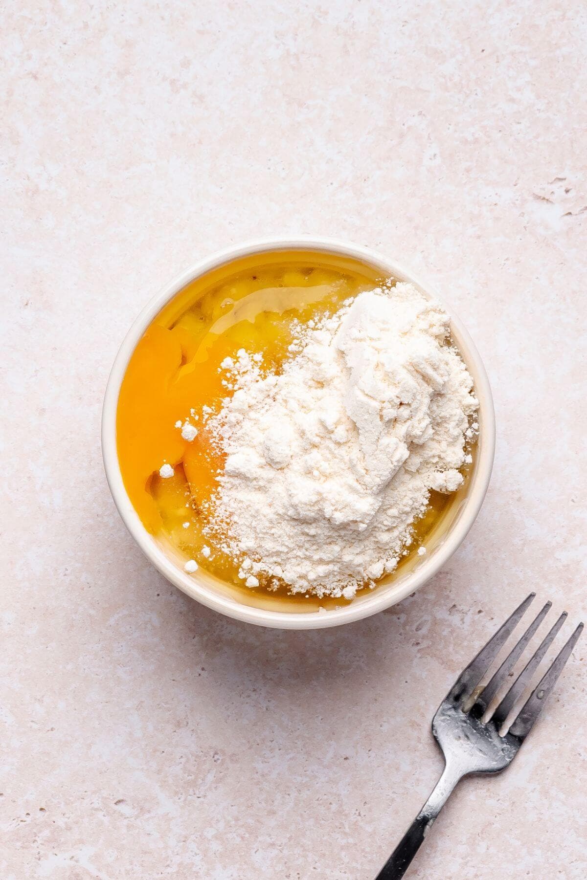 A bowl containing cracked eggs and a pile of flour on top, with a metal fork placed beside the bowl on a light-colored surface.