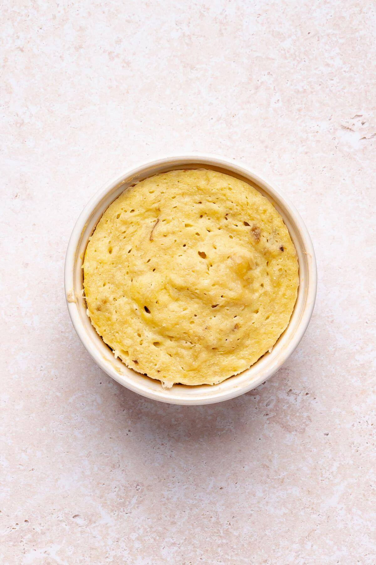 A round, golden mug cake sits in a white ramekin on a light beige surface, pictured from above.