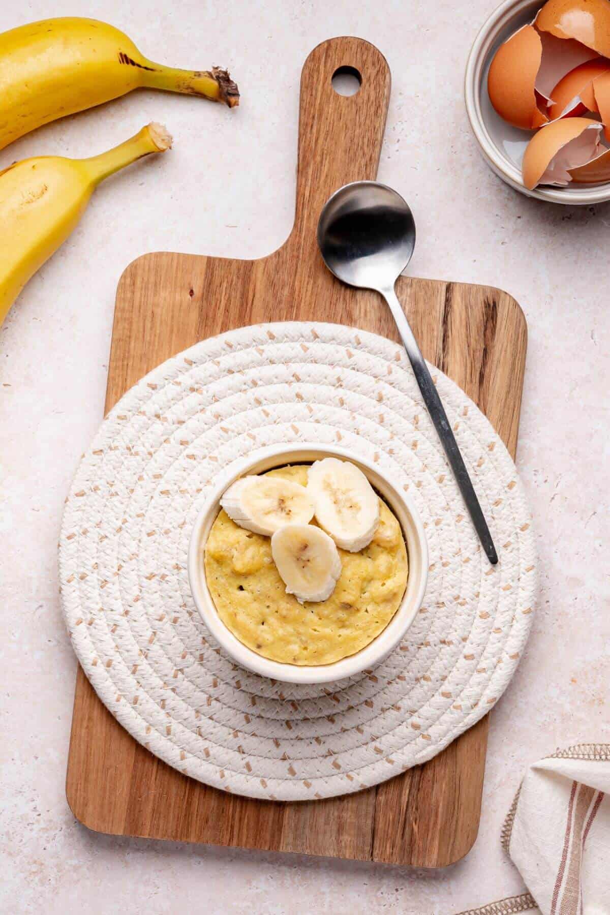 A bowl of banana oatmeal topped with banana slices sits on a woven mat and wooden board, with a spoon, banana, and eggshells nearby.