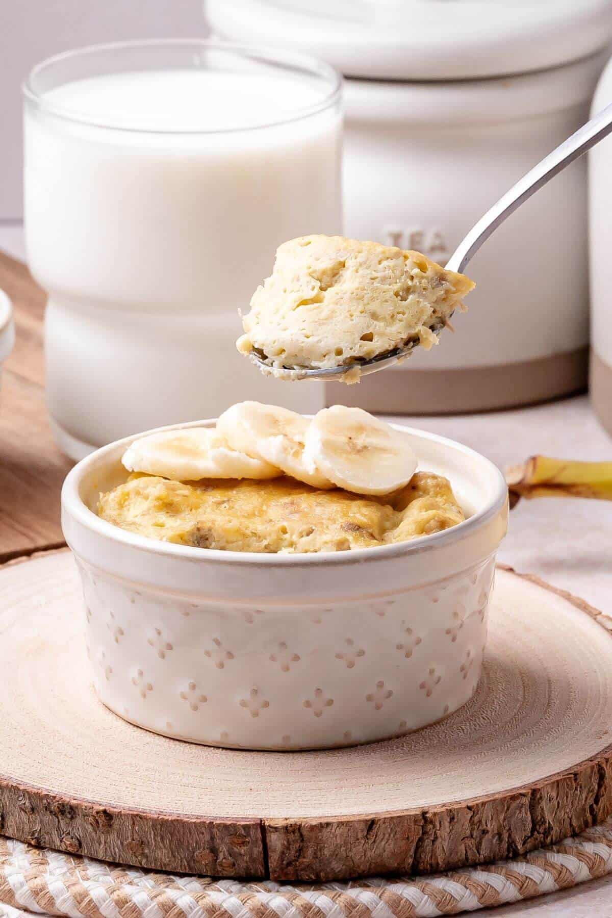 A white ramekin filled with a baked dish topped with banana slices, with a spoon lifting a portion. Glass of milk and containers are in the background.