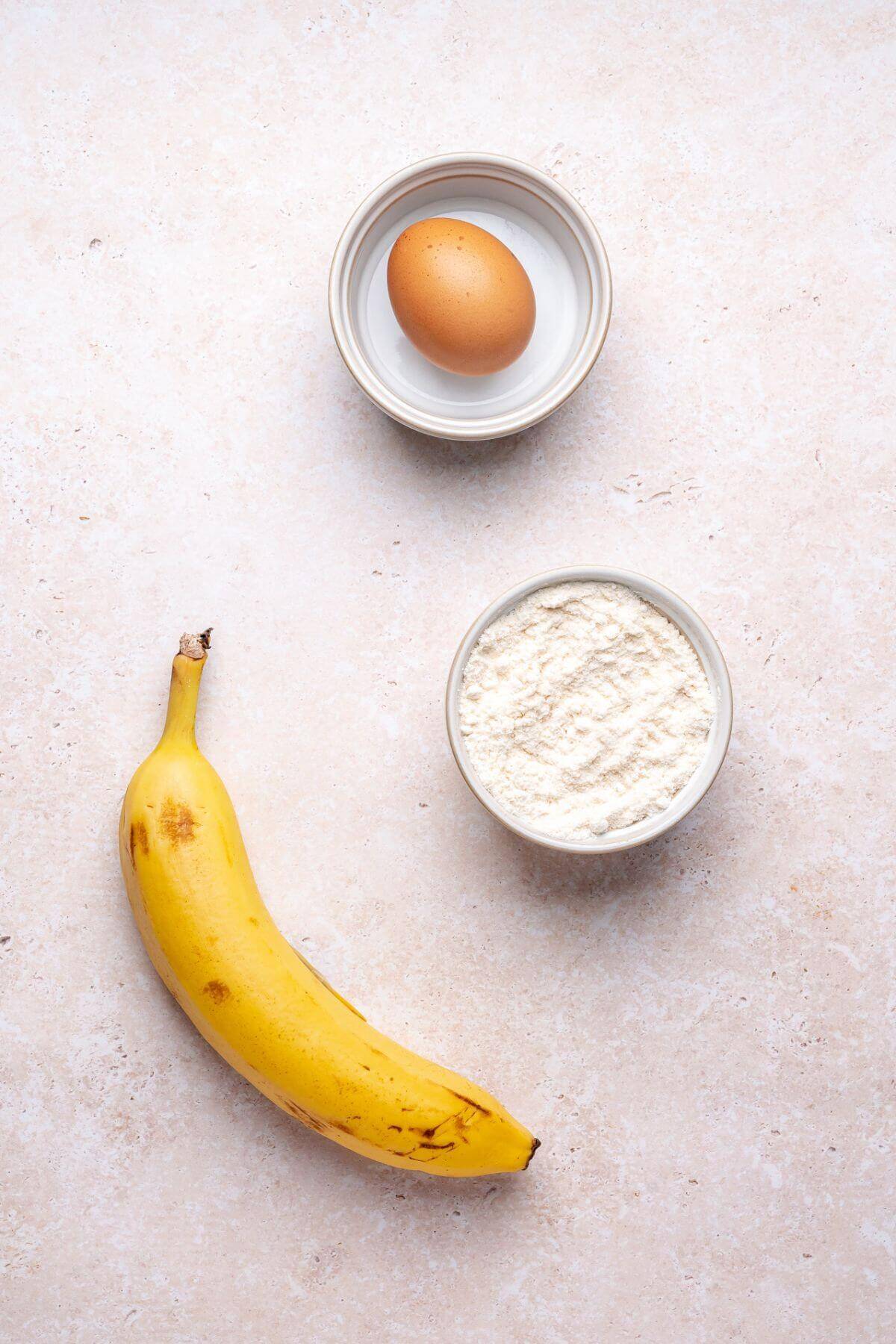 A whole banana, a bowl with a raw egg, and a bowl of flour are arranged on a light-colored surface.