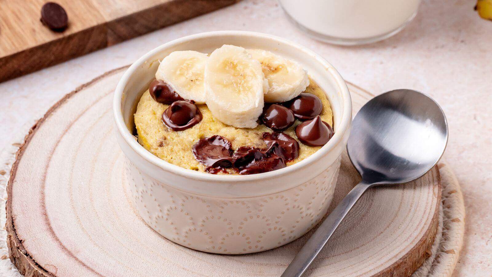 A ceramic ramekin filled with a baked muffin topped with banana slices and chocolate chips sits on a wooden coaster beside a metal spoon.