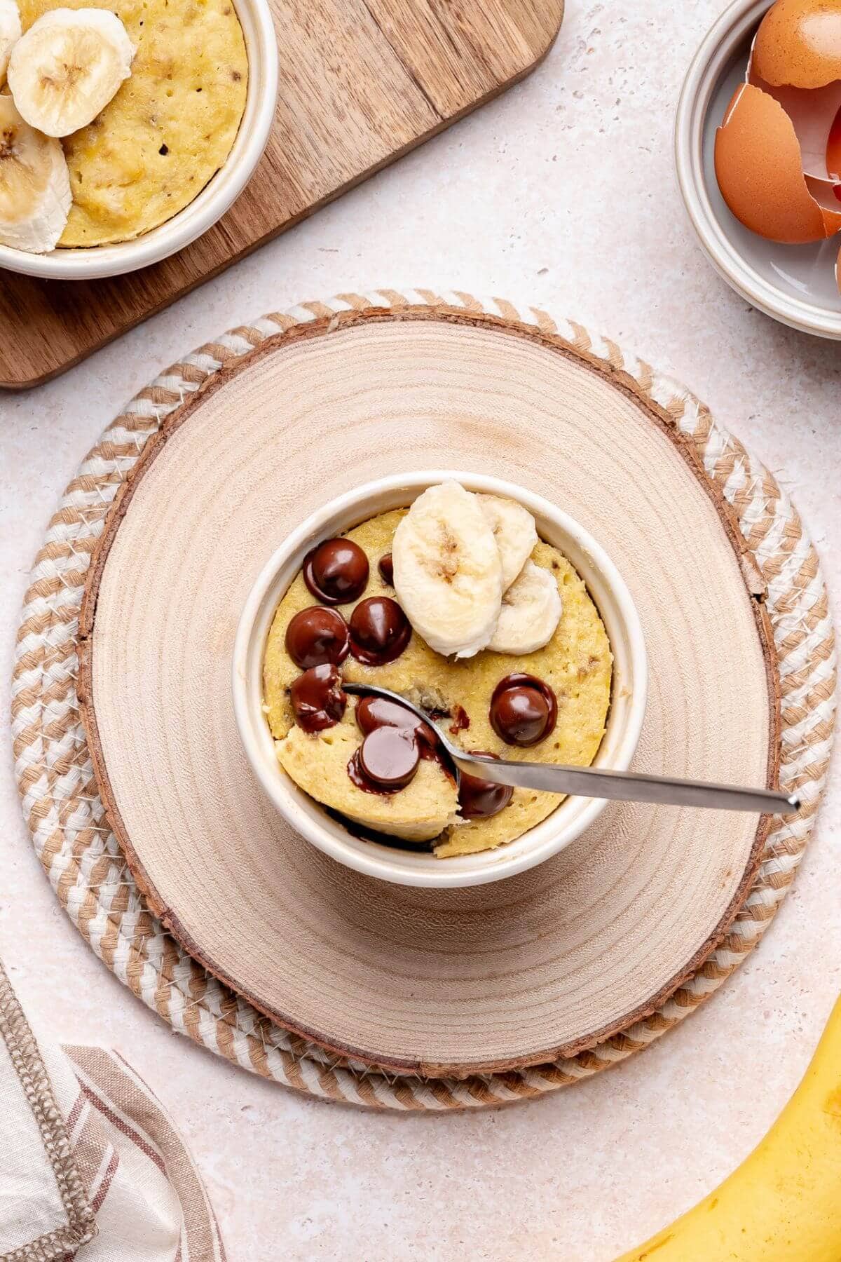 A mug cake topped with banana slices and chocolate chips sits on a wooden placemat, with a spoon in the mug and eggshells, banana slices, and a napkin nearby.