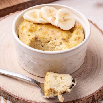 A small ramekin filled with banana mug cake, topped with banana slices. A spoon with a piece of the cake rests on the wooden surface beside it.
