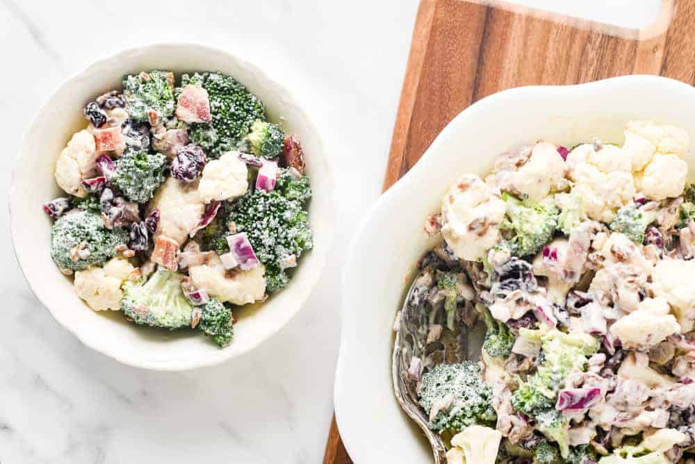 Two bowls of broccoli salad with cauliflower, red onion, dried cranberries, and creamy dressing on a marble surface.