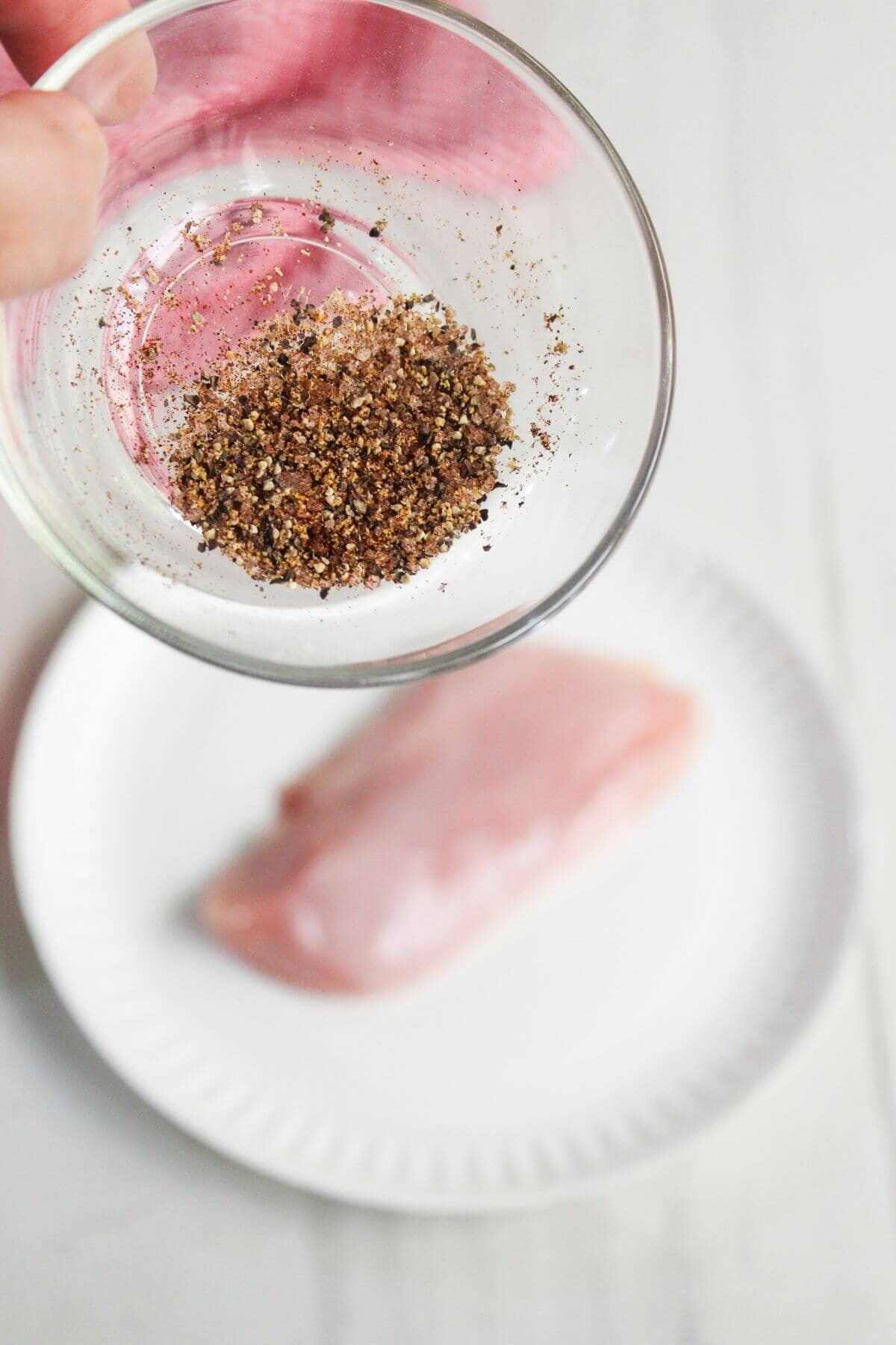 A hand holds a small bowl of mixed spices above a raw chicken breast on a white plate.