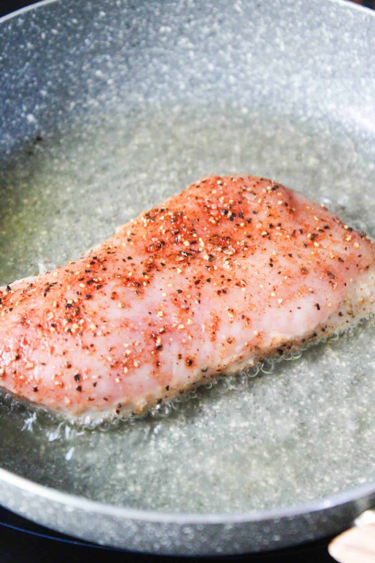 A seasoned piece of meat is frying in oil in a pan, with visible bubbling around the edges.