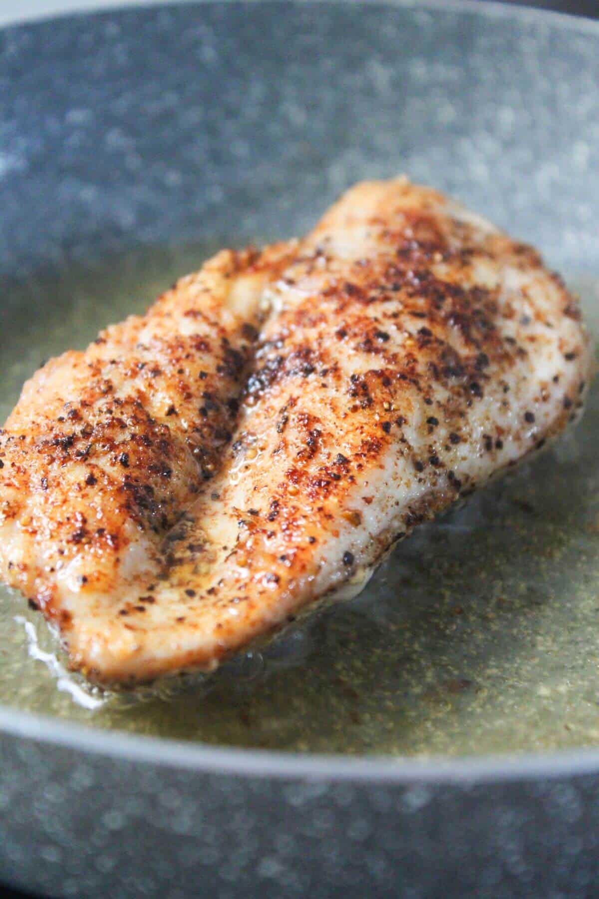 A seasoned piece of chicken breast is being pan-fried in oil in a skillet.