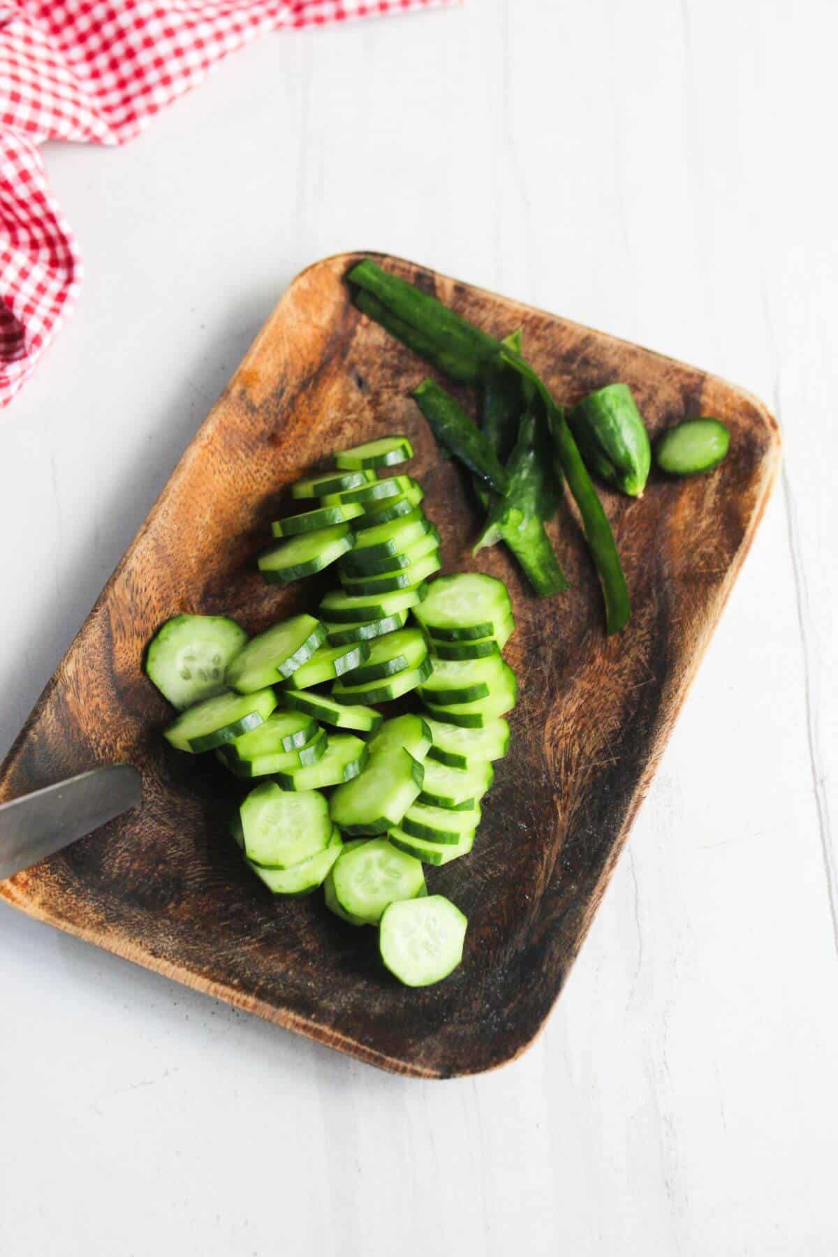 A wooden tray with cucumber slices and cucumber ends on a white surface, next to a red and white checkered cloth.