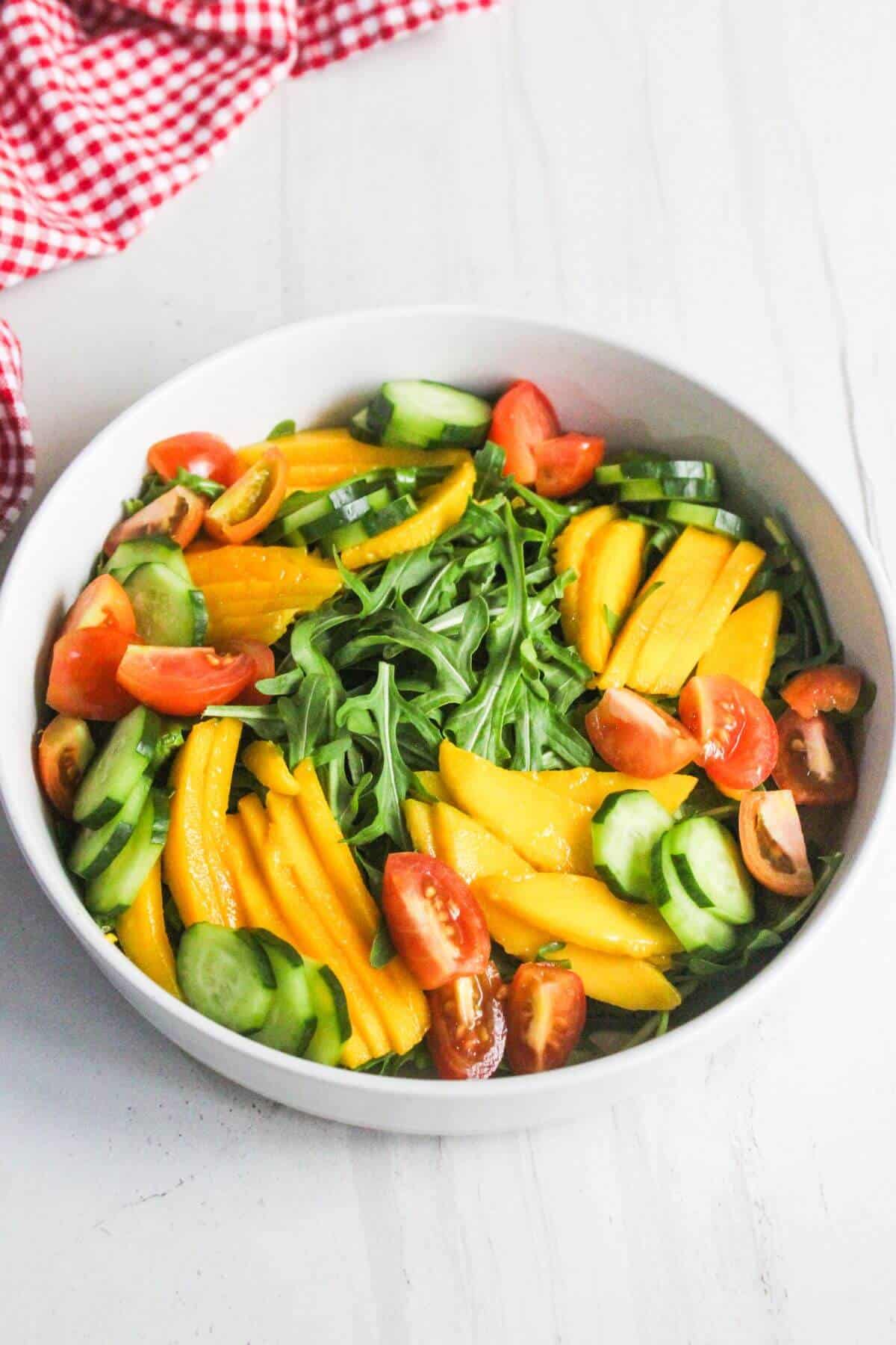A white bowl with arugula, sliced mango, cucumber, and tomato wedges on a white surface with a red checkered cloth in the background.