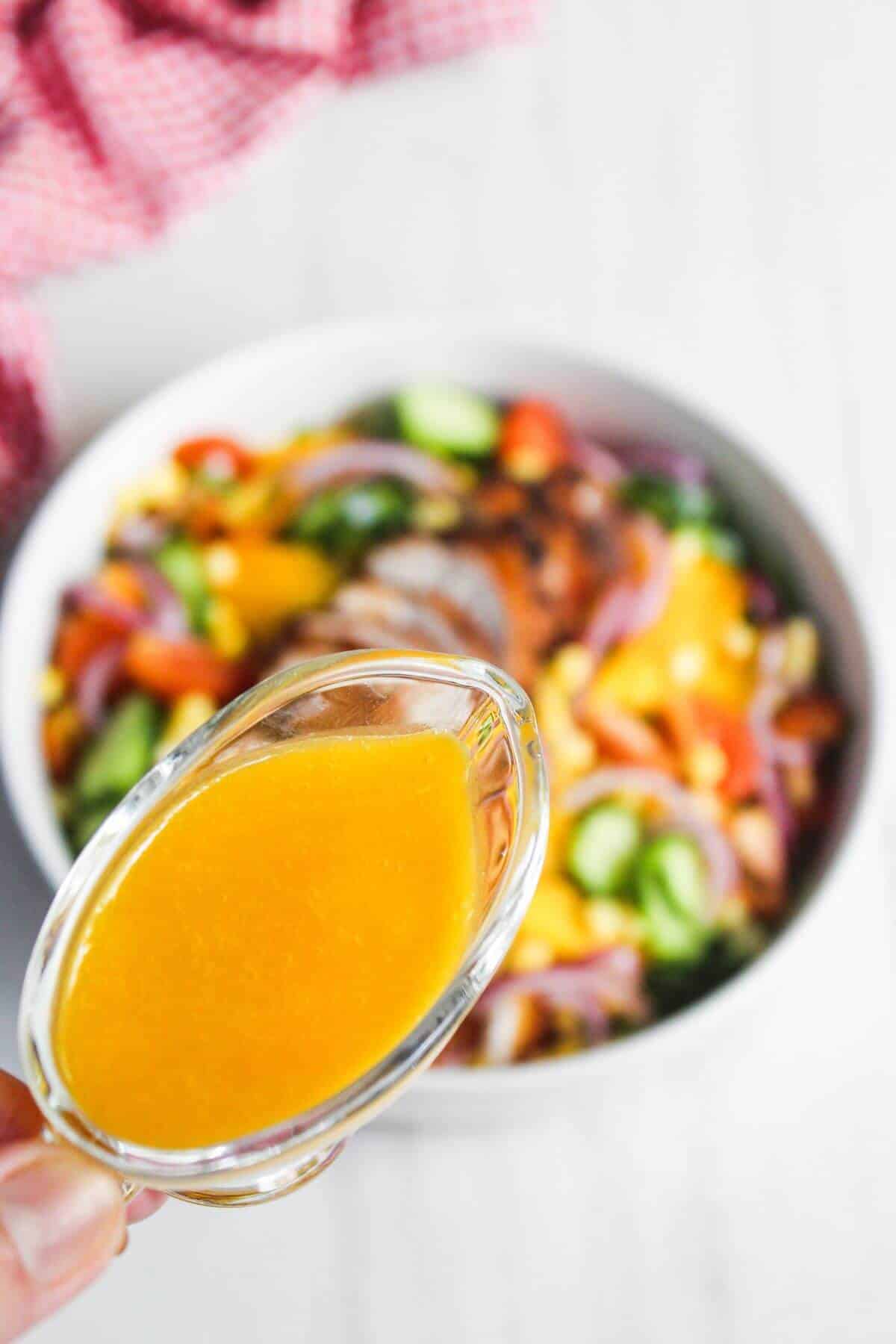 A hand holds a small glass container of orange salad dressing above a bowl of colorful mixed salad.
