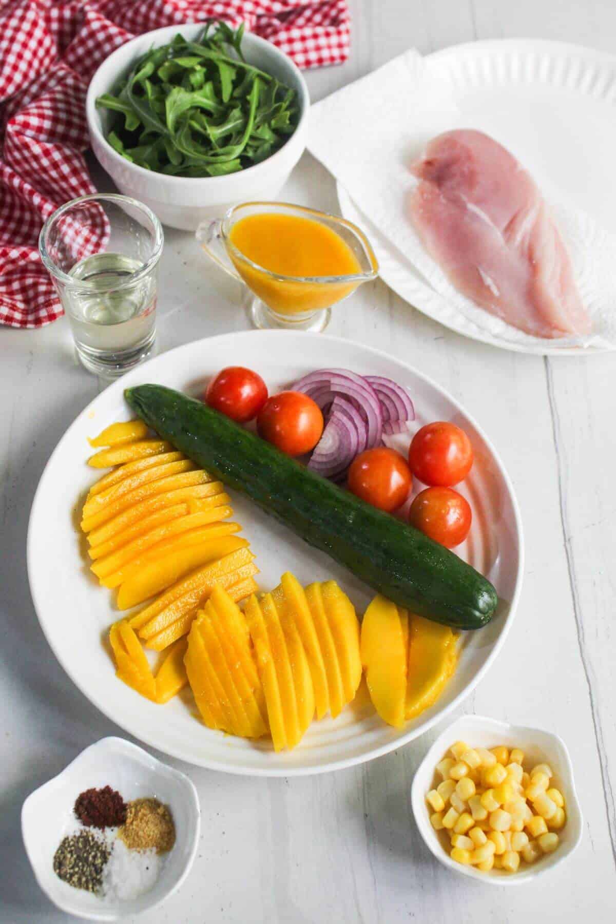 A plate with sliced mango, cucumber, cherry tomatoes, and red onion, surrounded by bowls of arugula, corn, spices, fish fillets, a glass of liquid, and a small jug of dressing.
