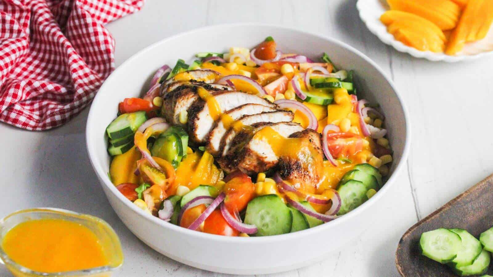 A bowl of salad with grilled chicken slices, cucumbers, tomatoes, red onions, corn, and mango slices, served with a side of yellow dressing.