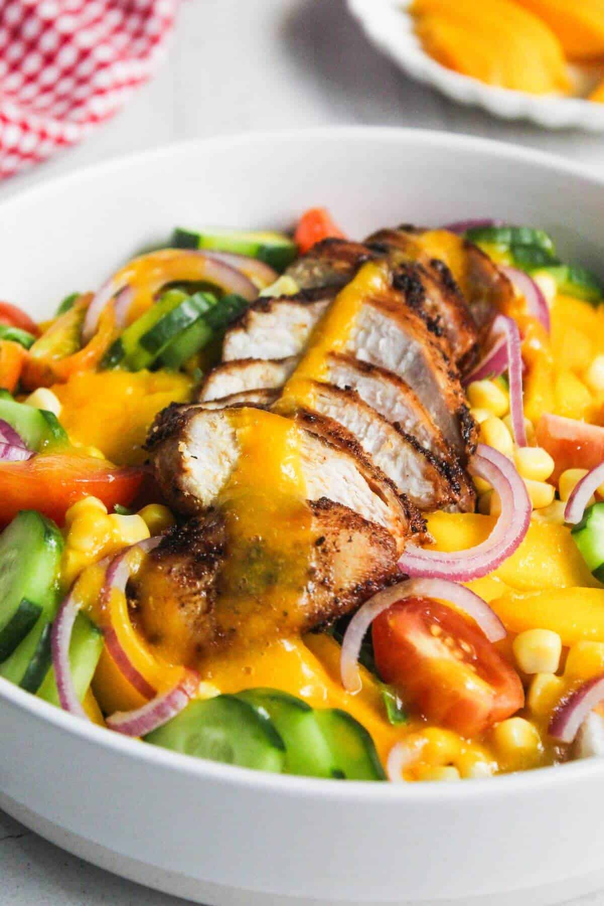 A bowl of salad with sliced grilled chicken breast, cherry tomatoes, cucumber, corn, red onion, and a yellow dressing on top.