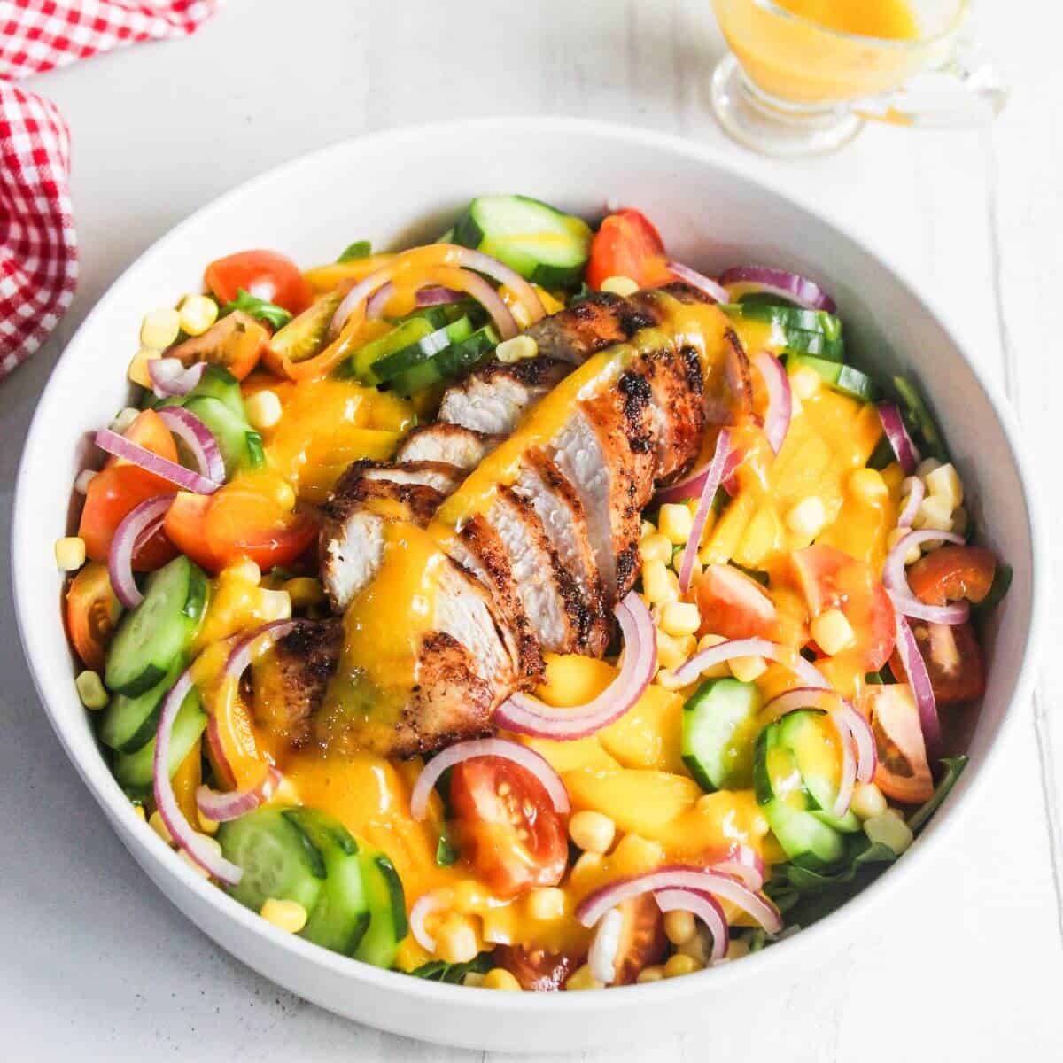 A bowl of salad with sliced grilled chicken, cucumbers, cherry tomatoes, red onions, corn, and yellow dressing on top.