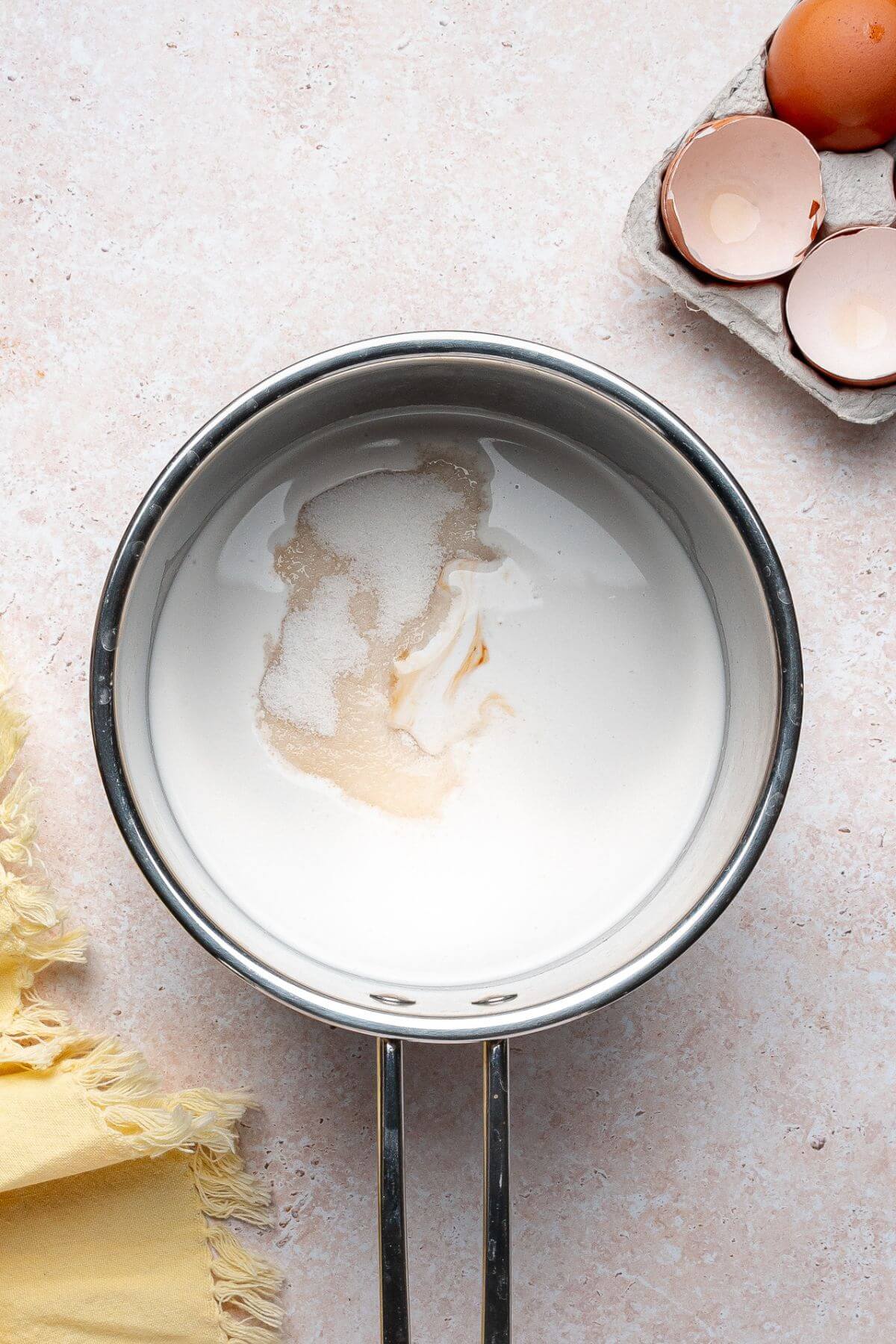 A saucepan containing milk, sugar, and vanilla sits on a light countertop next to a carton of cracked eggs and a yellow cloth.
