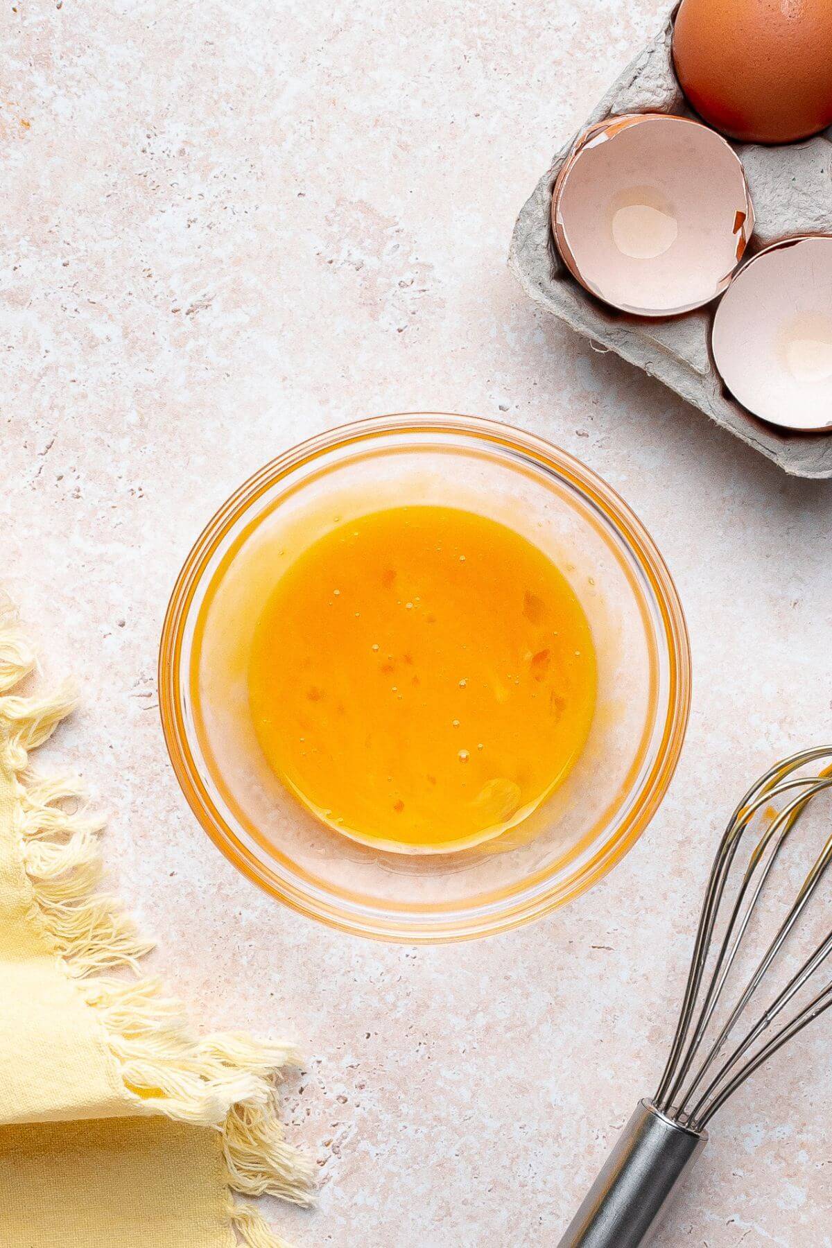 A glass bowl with beaten eggs sits on a light countertop. Next to it are a whisk, a yellow cloth, and an egg carton with empty eggshells.
