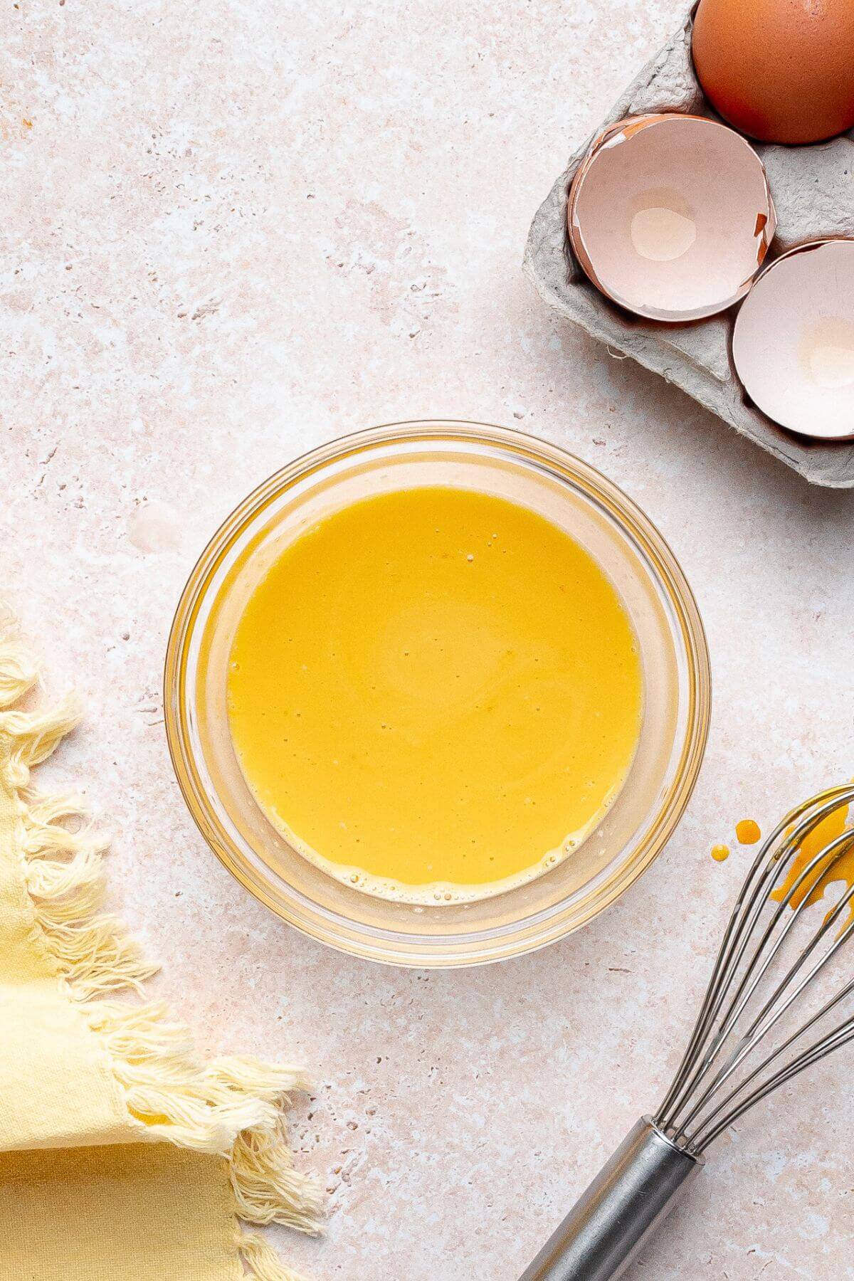 A glass bowl filled with beaten eggs sits on a light countertop near a metal whisk, egg shells in a carton, and a yellow cloth.