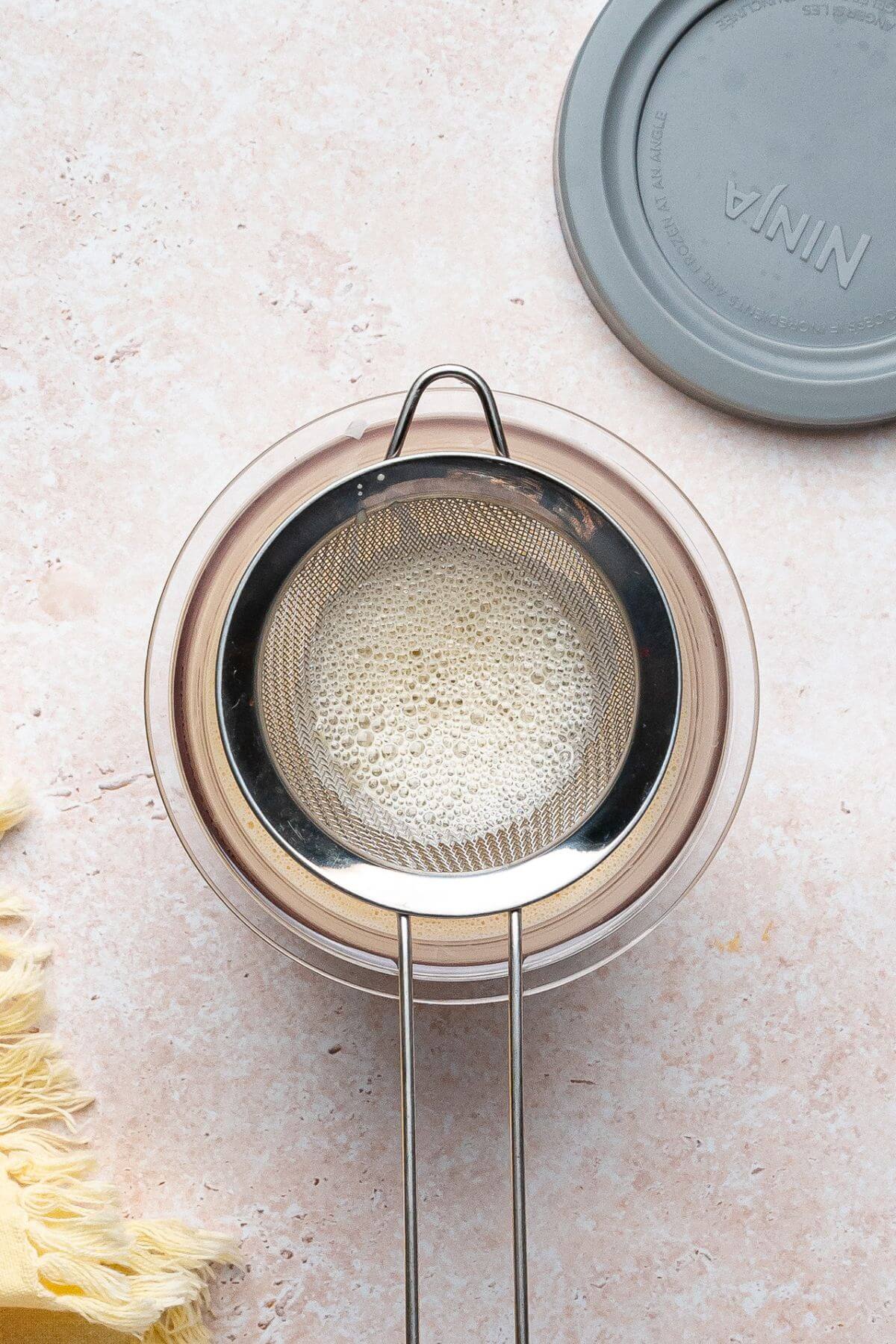 A metal strainer sits over a clear bowl, containing foamy liquid, with a gray Ninja lid and yellow cloth nearby on a light pink surface.